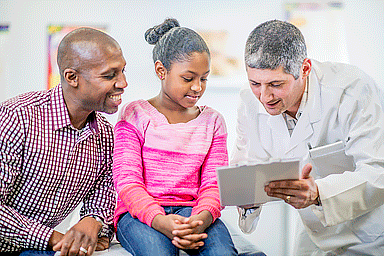 A doctor is talking to his patient and her father. The little girl is sitting on the examination table and is looking at the chart with her doctor.