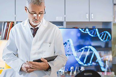 Male genetic research scientist working with a tablet in a laboratory with a computer monitor in the background showing DNA structure.