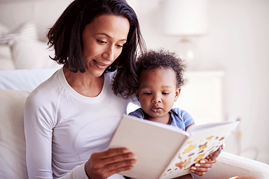 Close up of mixed race young adult mother sitting in an armchair reading a book with her baby son